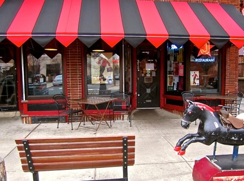 A storefront with a red and black awning, featuring a bench and a vintage horse ride-on toy outside.