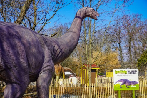 A large dinosaur statue stands in a park, with trees and a sign that reads "The Journey Continues" in the background.