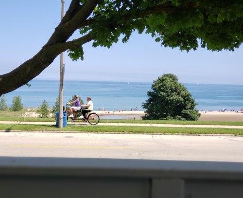 A couple rides a bicycle along a scenic beachside path with a view of the lake and distant sailboats.