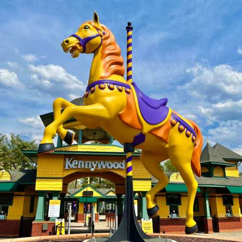 A vibrant yellow carousel horse stands in front of the Kennywood amusement park entrance under a blue sky.