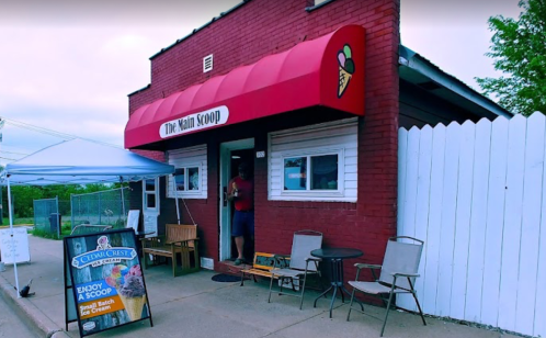A red-brick ice cream shop with a sign reading "The Main Scoop," featuring outdoor seating and a colorful awning.