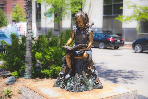 A bronze statue of a girl sitting on a rock, reading a book, surrounded by greenery and urban scenery.