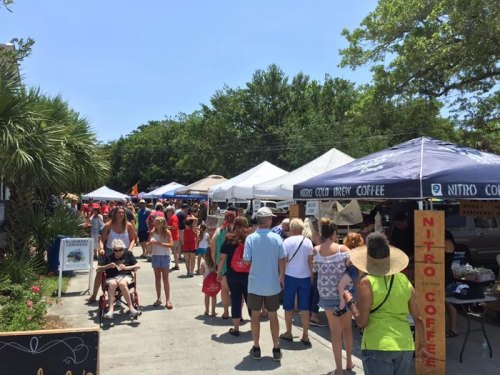 A bustling outdoor market scene with people browsing tents and stalls under a clear blue sky.