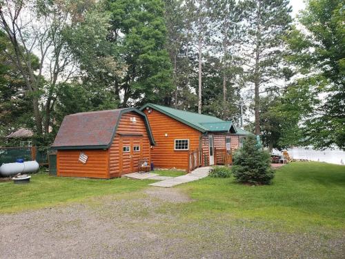A cozy log cabin surrounded by trees, with a green roof and a pathway leading to a lake in the background.