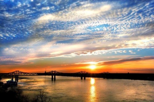 A vibrant sunset over a river, with colorful clouds and a bridge silhouetted against the sky.
