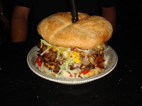 A large, stacked burger with lettuce, tomatoes, cheese, and onions on a plate, topped with a black straw.