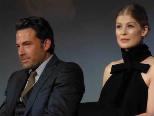 A man in a suit and a woman in a black dress sit side by side, both looking thoughtfully at the audience.