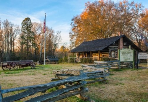 Historic log cabin with a wooden fence, flagpole, and sign, set against a backdrop of trees and a sunset sky.