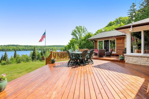 A wooden deck with a dining set overlooks a lake and lush greenery, featuring an American flag in the background.