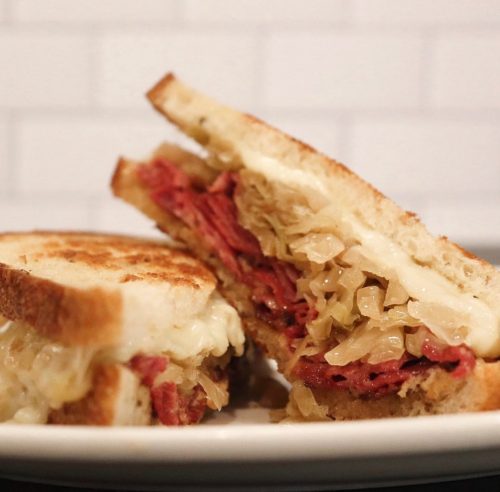 A close-up of a grilled sandwich with layers of corned beef, sauerkraut, and melted cheese on toasted bread.
