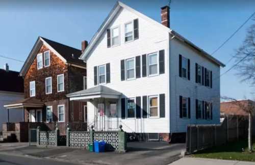 A two-story white house with black shutters, next to a brown shingle house, on a residential street.