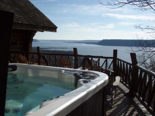 A hot tub on a wooden deck overlooking a serene lake and distant hills under a clear blue sky.