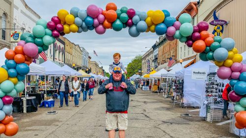 A man carries a boy on his shoulders under a colorful balloon arch at a street fair with vendors and attendees.
