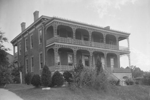 Historic brick building with ornate ironwork balconies, surrounded by greenery and a clear sky.
