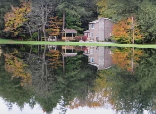 A serene lake reflects a house and colorful autumn trees, surrounded by lush greenery.