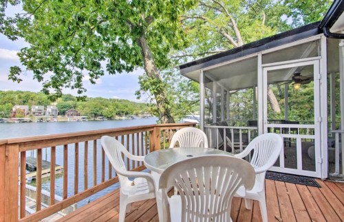 A wooden deck with a table and chairs overlooks a serene lake surrounded by trees and houses.