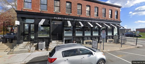 Exterior of a plant-based food hall and marketplace, featuring large windows and a striped awning. A parked car is visible.