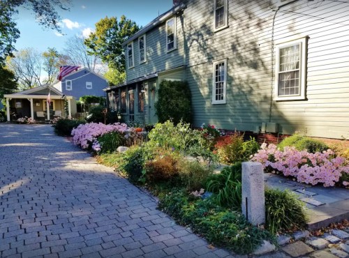 A charming house with a stone pathway, surrounded by colorful flower beds and greenery on a sunny day.