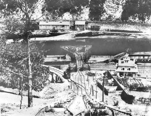 Black and white photo of a mining area with buildings, tracks, and a bridge over a body of water.