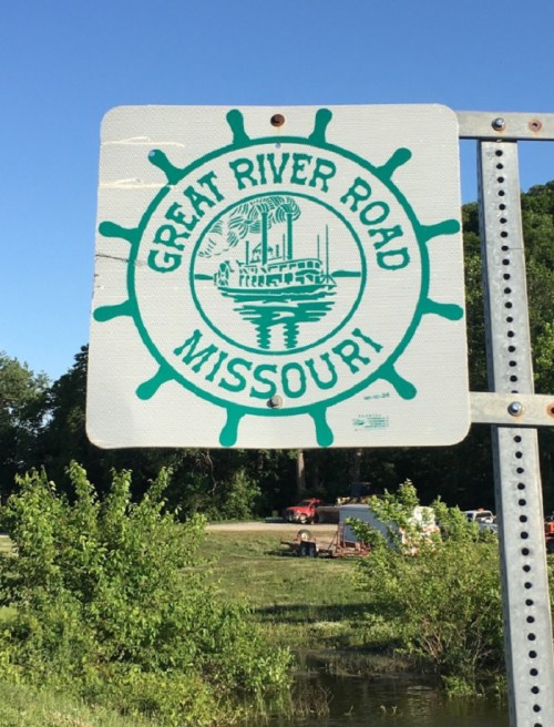 Sign for Great River Road in Missouri, featuring a steamboat graphic and surrounded by a circular design.