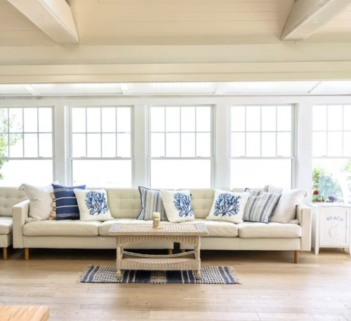 Bright living room with a white sofa, blue and white pillows, and a woven coffee table by large windows.