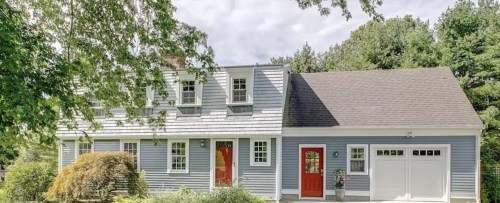 A charming blue house with a red door, surrounded by greenery and trees under a cloudy sky.