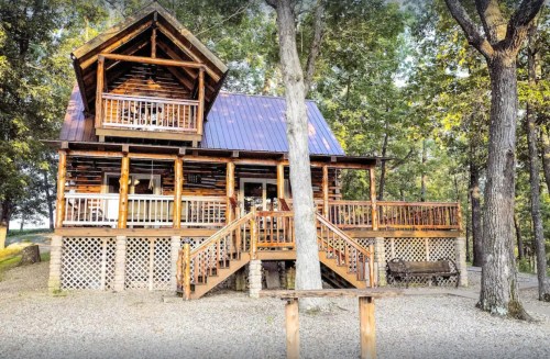 A rustic wooden cabin with a blue roof, surrounded by trees and featuring a spacious deck and stairs.