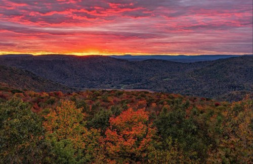 Vibrant sunset over rolling hills, with colorful autumn foliage in the foreground and dramatic clouds in the sky.