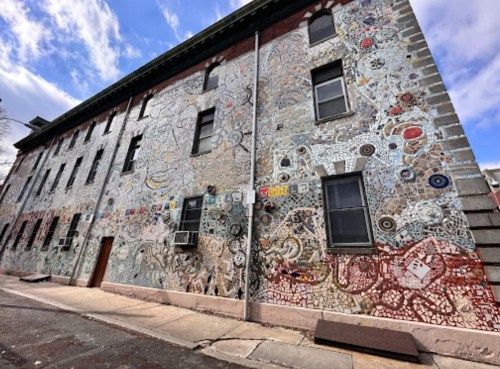 Colorful mosaic-covered wall of a building, featuring intricate patterns and designs under a partly cloudy sky.