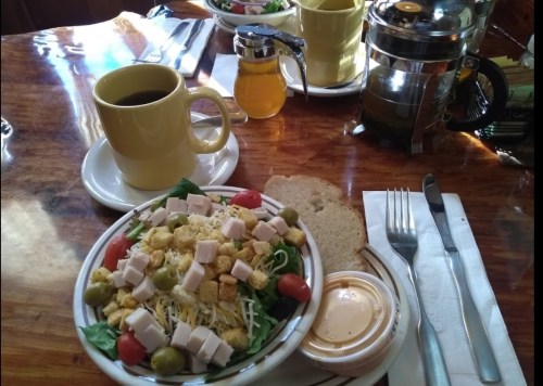A salad with turkey, tomatoes, and olives, alongside a cup of coffee and a small jar of honey on a wooden table.