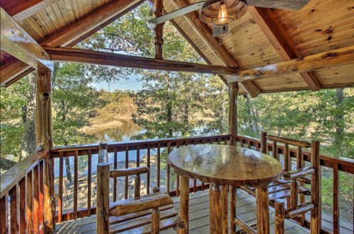 Cozy wooden balcony with a table and chairs overlooking a serene river and lush trees.