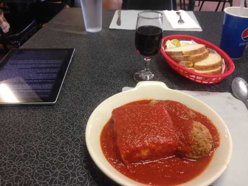 A plate of meatloaf with tomato sauce, a glass of red wine, a basket of bread, and an iPad on a table.