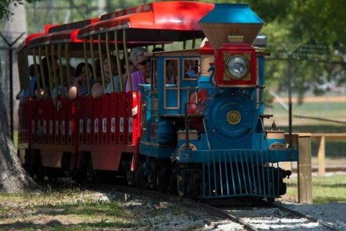 A colorful miniature train with passengers rides along a track in a park, surrounded by trees.