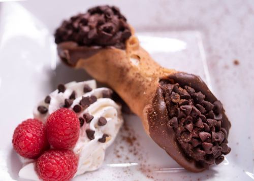 A chocolate-dipped cannoli filled with mini chocolate chips, served with whipped cream and fresh raspberries.