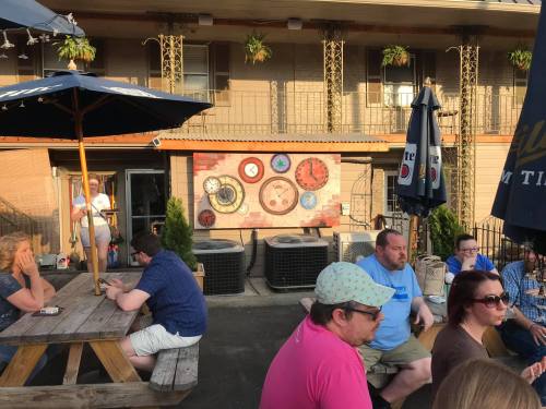 A lively outdoor scene at a bar with people sitting at picnic tables and decorative wall art in the background.