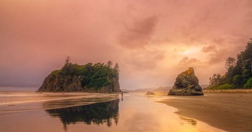 A serene beach at sunset, featuring rocky formations and lush greenery, with soft clouds reflecting in the water.