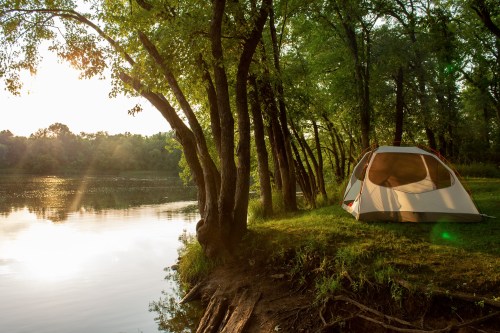 A tent by a serene river, surrounded by trees, with sunlight filtering through the leaves at sunset.