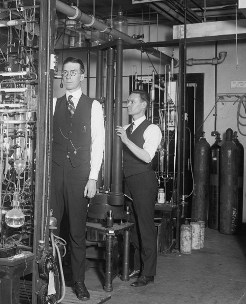 Two men in vintage suits work in a laboratory filled with glass equipment and gas cylinders.