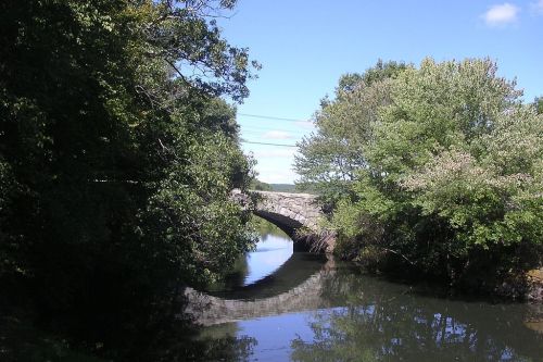 A stone bridge arches over a calm river, surrounded by lush green trees under a clear blue sky.