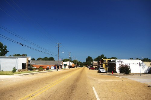 A quiet street lined with small businesses under a clear blue sky, with power lines and trees in the background.