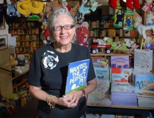 An elderly woman smiles while holding a children's book titled "Wanted: The Perfect Pet" in a cozy bookstore.