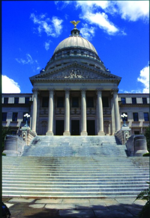 A grand building with a large dome and columns, featuring a statue on top, set against a blue sky with clouds.