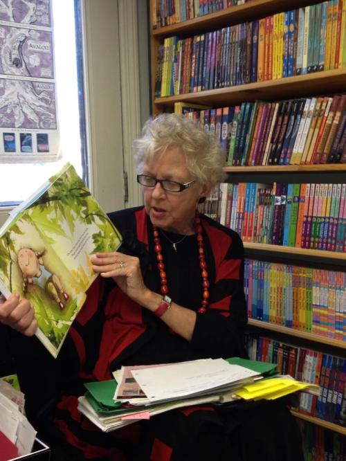 A woman with curly hair and glasses reads a children's book, surrounded by colorful bookshelves.