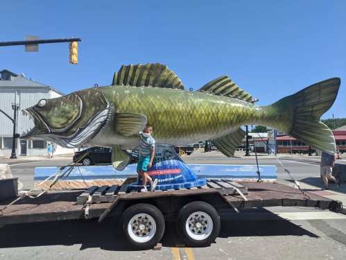 A person stands next to a large, colorful fish statue on a trailer in a sunny outdoor setting.