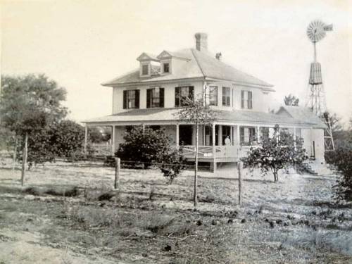 Historic black-and-white photo of a large house with a porch, surrounded by trees and a windmill in the background.