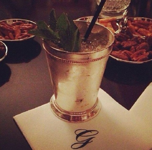 A silver cup filled with a cocktail garnished with mint, placed on a napkin with a "G" logo, surrounded by snacks.