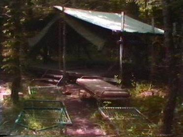 A makeshift tent in a forest, with a cot and several metal frames scattered around in the grass.