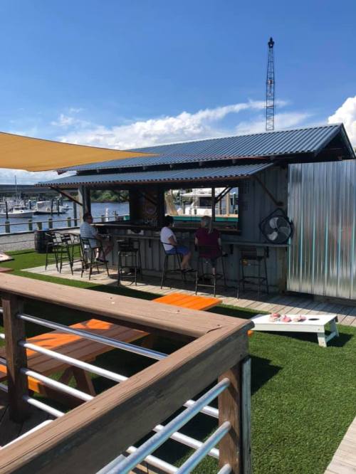 A waterfront bar with patrons sitting outside, surrounded by green grass and a clear blue sky.