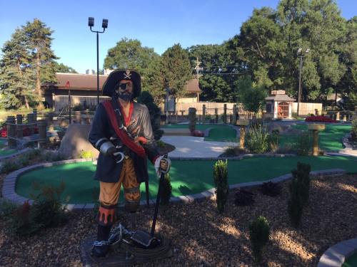 A pirate statue stands in a mini-golf course surrounded by green turf and landscaped gardens.