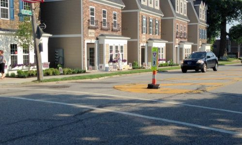 A street scene featuring houses, a crosswalk sign, and a car driving past on a sunny day.